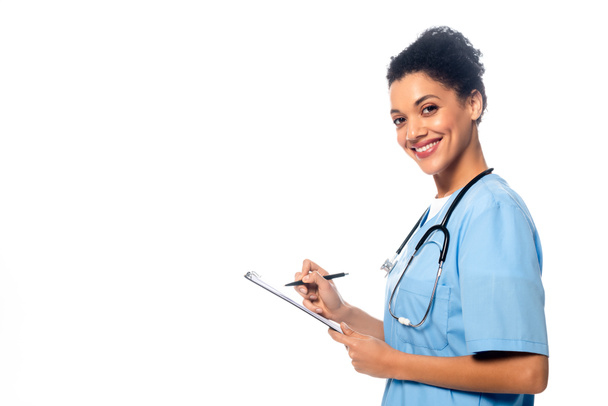 Zijaanzicht van Afrikaanse Amerikaanse verpleegster met stethoscoop schrijven op klembord en glimlachen geïsoleerd op wit - Foto, afbeelding