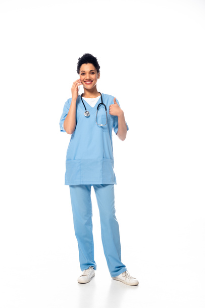 Full length view of happy african american nurse with thumb up, stethoscope and smartphone on white - Photo, Image