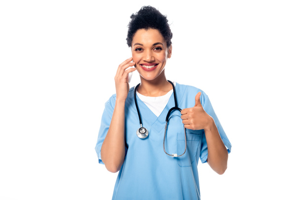 Вид спереди счастливой африканской медсестры с большим пальцем вверх, стетоскопом и смартфоном, изолированным на белом
 - Фото, изображение