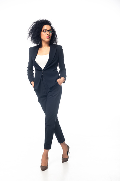 Full length view of confident african american businesswoman walking with hands in pockets on white background - Photo, Image
