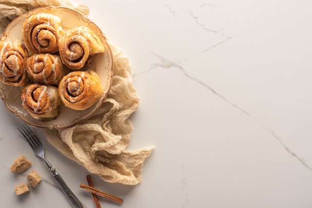 bovenaanzicht van verse zelfgemaakte kaneelbroodjes op marmeren oppervlak met bruine suiker, kaneelstokjes, vork en doek - Foto, afbeelding