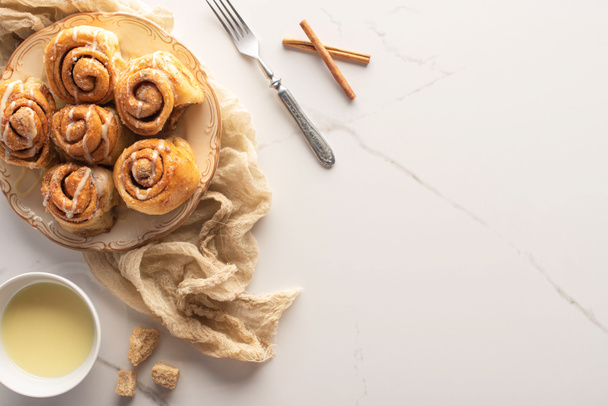 top view of fresh homemade cinnamon rolls on marble surface with condensed milk, brown sugar, cinnamon sticks, fork and cloth - Photo, Image