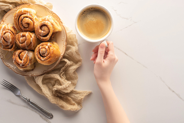 vista recortada de la mujer sosteniendo taza de café cerca de rollos de canela caseros en la superficie de mármol con tenedor y tela
 - Foto, Imagen