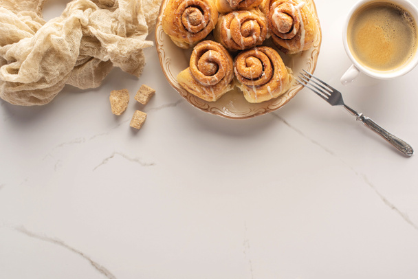 top view of fresh homemade cinnamon rolls on marble surface with cup of coffee, fork and cloth - 写真・画像