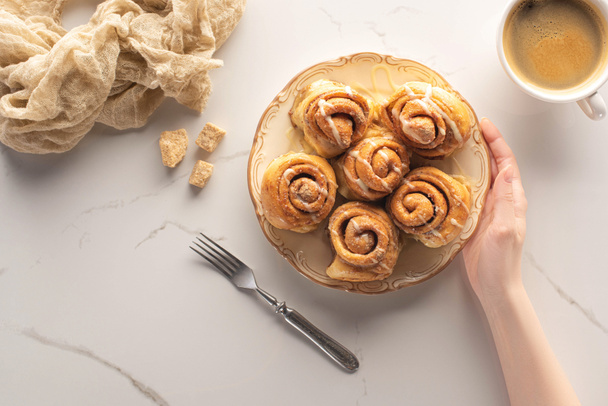 cropped view of woman holding homemade cinnamon rolls on marble surface with fork, cup of coffee and cloth - Photo, Image