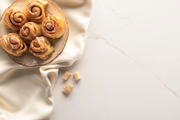 bovenaanzicht van verse zelfgemaakte kaneelbroodjes op zijden doek op marmeren oppervlak met bruine suiker - Foto, afbeelding