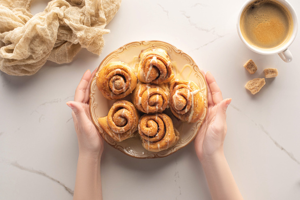 cropped view of woman holding plate with fresh homemade cinnamon rolls on marble surface with cup of coffee and cloth - Photo, Image