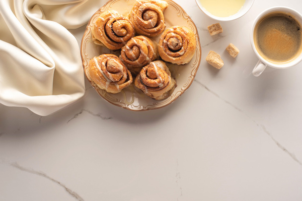 top view of fresh homemade cinnamon rolls on marble surface with cup of coffee, brown sugar, condensed milk and silk cloth - Φωτογραφία, εικόνα