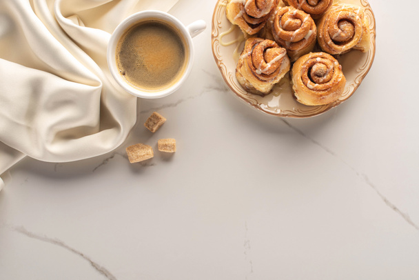 Taze ev yapımı tarçın rulolarının mermer yüzeyinde bir fincan kahve, kahverengi şeker ve ipek kumaşla üst görüntüsü - Fotoğraf, Görsel