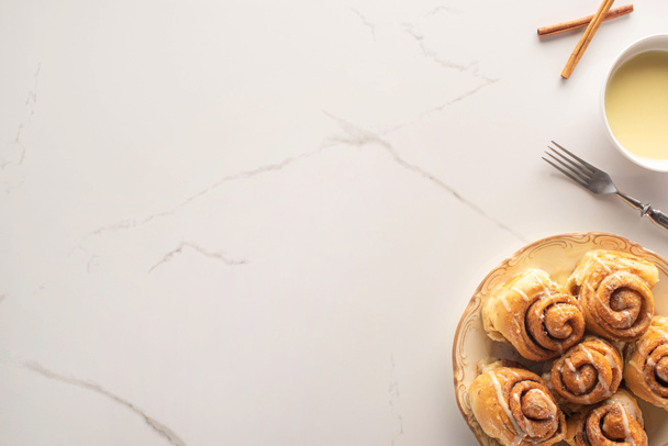 top view of fresh homemade cinnamon rolls on marble surface with condensed milk, fork - Photo, Image