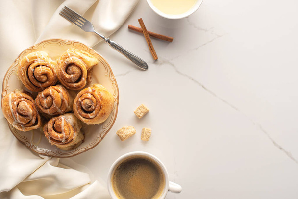 top view of fresh homemade cinnamon rolls on marble surface with coffee, condensed milk, fork and satin cloth - Photo, Image