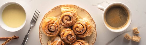 top view of fresh homemade cinnamon rolls on marble surface with cup of coffee, fork and condensed milk, panoramic shot - Photo, Image