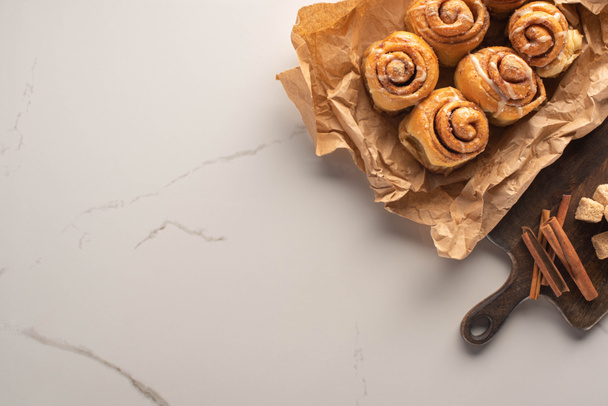 top view of fresh homemade cinnamon rolls on parchment paper on marble surface with cutting board with brown sugar and cinnamon sticks - Photo, Image