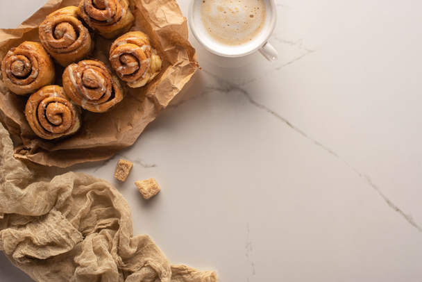 vista superior de rollos de canela caseros frescos en papel pergamino sobre superficie de mármol con taza de café, azúcar morena y tela
 - Foto, Imagen