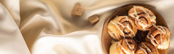 vrchní pohled na čerstvé domácí skořicové rolky na hedvábné bílé látce s hnědým cukrem, panoramatický záběr - Fotografie, Obrázek