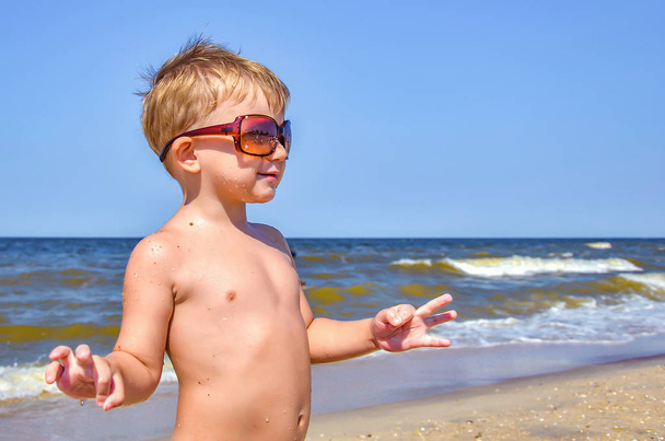 Ein fröhlicher Junge mit Sonnenbrille steht am Strand, die Arme ausgebreitet. Kinder am Strand entspannen. - Foto, Bild