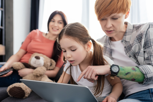 Selective focus of mother using laptop with daughter near smiling parent with teddy bear on couch - Foto, imagen