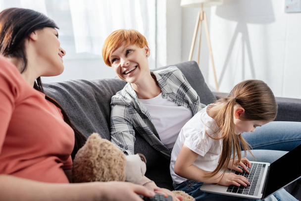 Selective focus of smiling same sex couple looking at each other near child with laptop on couch - Фото, изображение
