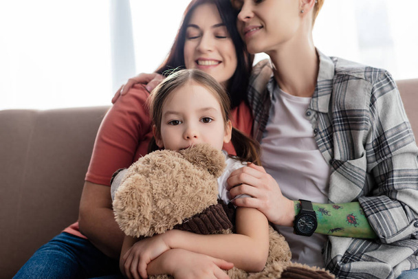 Kind mit Teddybär blickt auf Couch in die Kamera und lächelt gleichgeschlechtliche Eltern an  - Foto, Bild