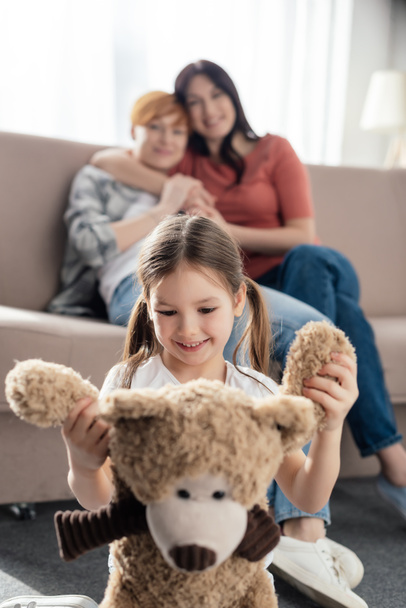 Selectieve focus van lachend kind spelen met teddybeer in de buurt van moeders omarmen op de bank  - Foto, afbeelding