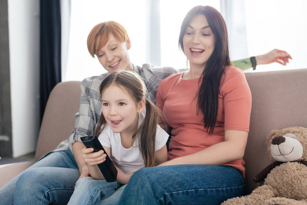 Cheerful kid with remote controller sitting near mothers on couch in living room - Photo, Image