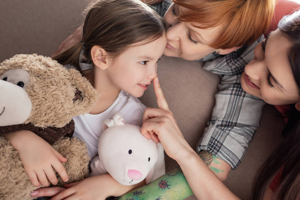 Smiling same sex parents kissing and touching nose of daughter with soft toys on couch  - Foto, imagen
