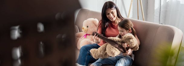 Выборочный фокус улыбающейся матери, смотрящей на дочь, играющую с плюшевым мишкой на диване, панорамный снимок
 - Фото, изображение