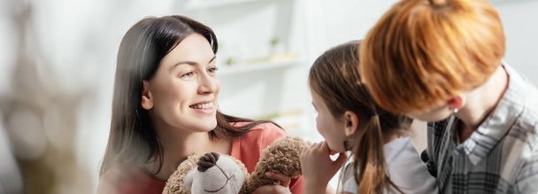 Selective focus of smiling mother holding teddy bear near daughter and parent in living room, panoramic shot - Фото, изображение
