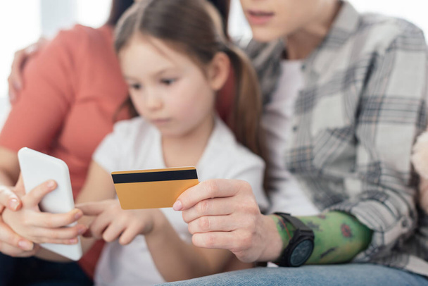 Selective focus of same sex parents holding credit card while daughter using smartphone at home  - Photo, Image
