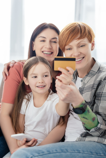 Selective focus of smiling kid with smartphone holding credit card near same sex parents on sofa  - Fotografie, Obrázek