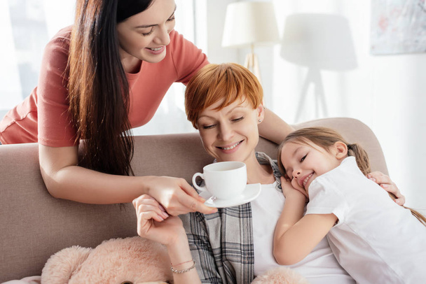 Smiling woman giving coffee to parent hugging happy daughter on sofa - Фото, изображение
