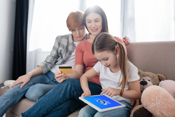 KYIV, UCRANIA - 10 DE FEBRERO DE 2020: Niño usando la tableta digital con la aplicación shazam en la pantalla cerca de padres del mismo sexo con tarjeta de crédito en el sofá en casa
 - Foto, Imagen