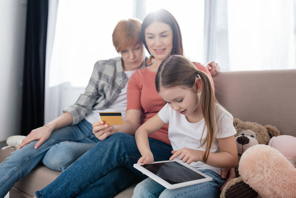 Ребенок с помощью цифрового планшета с чистым экраном рядом улыбающиеся однополые родители с кредитной картой на диване
 - Фото, изображение