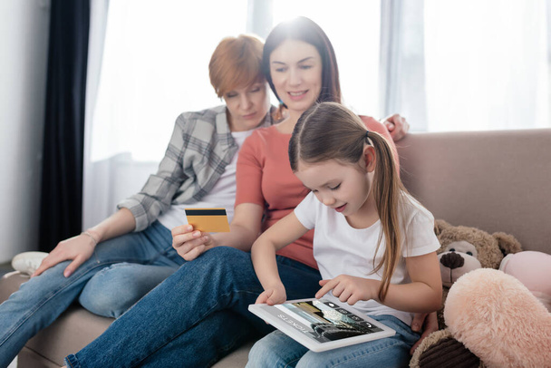 Kind nutzt digitales Tablet mit Ticket-App am Bildschirm in der Nähe gleichgeschlechtlicher Eltern mit Kreditkarte zu Hause - Foto, Bild