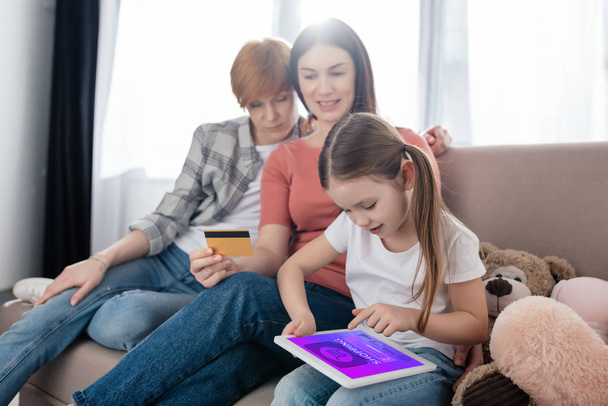 Child using digital tablet with shopping app on screen near same sex parents with credit card and soft toys on couch - Photo, Image