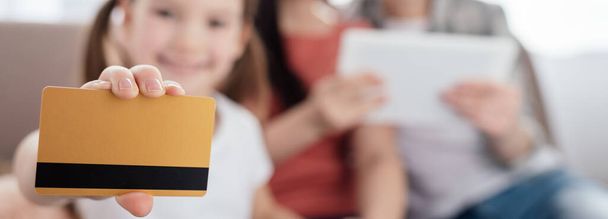 Foco seletivo de criança segurando cartão de crédito e sorrindo para a câmera perto de mães com tablet digital, tiro panorâmico
 - Foto, Imagem