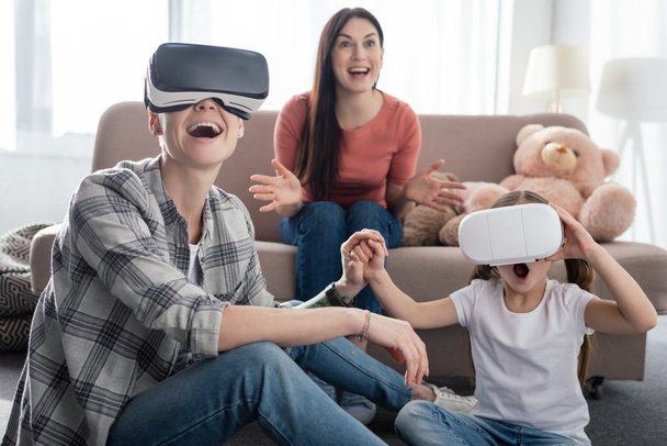 Same sex parents using virtual reality headsets with excited daughter in living room - Zdjęcie, obraz