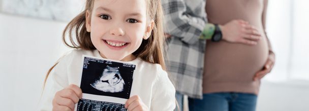 Выборочный фокус улыбающегося ребенка, держащего ультразвуковое сканирование ребенка рядом с матерью, обнимающей беременную женщину дома, панорамный снимок
 - Фото, изображение