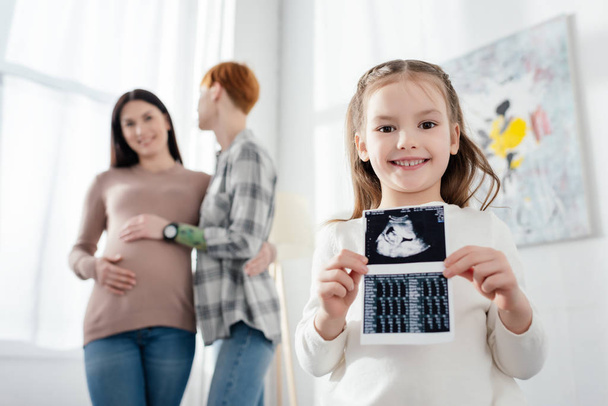Selective focus of smiling kid holding ultrasound scan of baby near woman embracing pregnant girlfriend  - Photo, Image