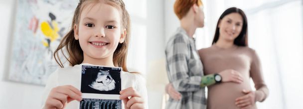 Bebek ultrasonu tutan gülümseyen çocuğun seçmeli odak noktası. Bu arada evde hamile bir kız arkadaşına sarılıyor. Panoramik çekim.  - Fotoğraf, Görsel