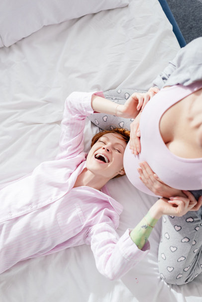 Top view of laughing woman in pajamas touching belly of pregnant girlfriend on bed  - Fotografie, Obrázek
