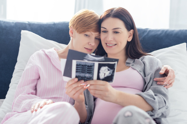 Selective focus of smiling same sex couple in pajamas looking at ultrasound scans of baby on bed  - Photo, Image