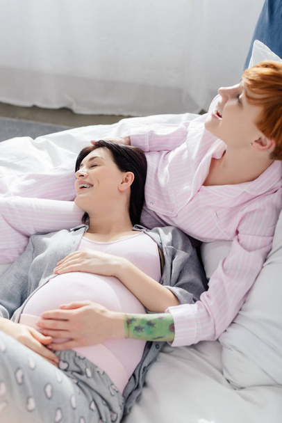 Smiling woman touching tummy of happy pregnant girlfriend on bed at home  - Foto, imagen
