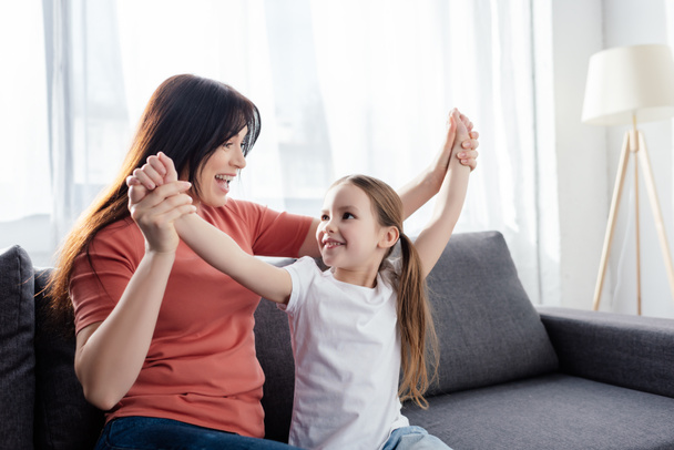 Happy mother playing with smiling daughter on couch at home  - Фото, изображение
