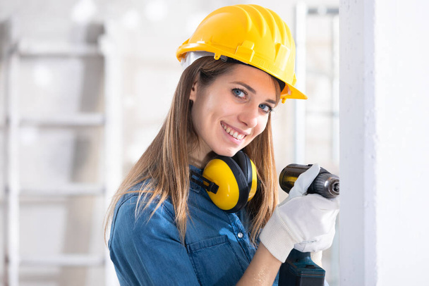 Nuori nainen Builder poraus reikä uusi valkoinen seinä päällä akkukäyttöinen käsi pora katsomassa, mitä hän tekee hymyillen
 - Valokuva, kuva