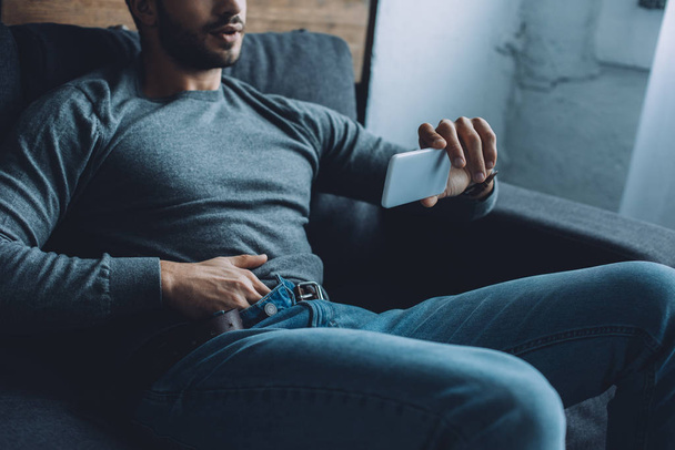 Cropped view of man masturbating while watching pornography on smartphone on sofa  - Fotó, kép