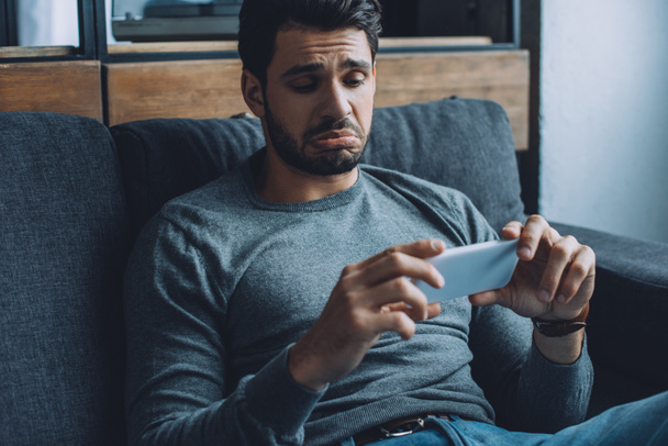 Trauriger Mann schaut Pornos auf Smartphone auf Couch im Wohnzimmer - Foto, Bild