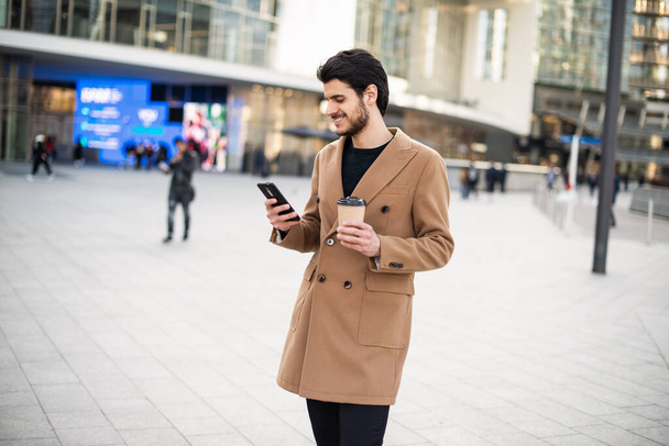 Man using his mobile phone and holding a cup of coffee while walking in a city square - Foto, Imagen