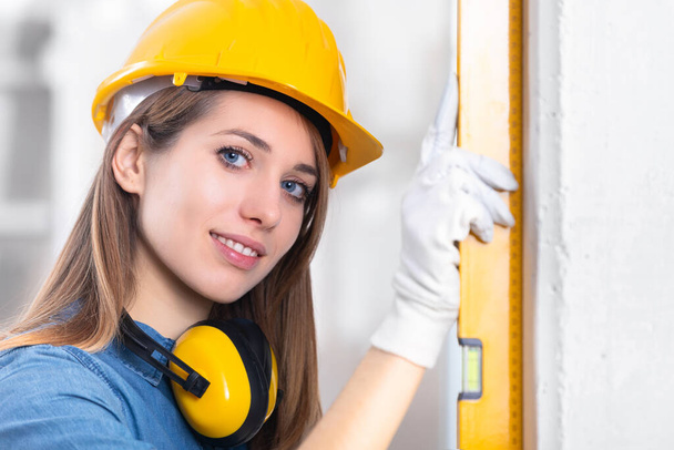 Attractive young female construction worker in yellow hardhat checking level of the wall with the spirit level and turning to smile at camera  - Φωτογραφία, εικόνα