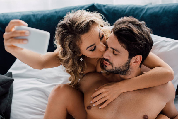 Selective focus of attractive woman kissing shirtless boyfriend while taking selfie with smartphone on bed  - Photo, Image
